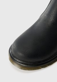 Bota de tornozelo em couro preto com bico arredondado, parte superior texturizada e sola de borracha grossa com sulcos horizontais perto da base.