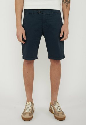 Uomo che indossa pantaloncini blu scuro fino al ginocchio, sneakers bianche e una maglietta bianca, in piedi con le mani rilassate ai lati.