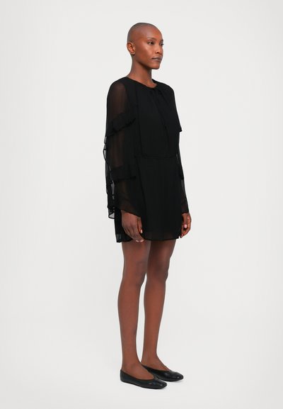 Black, sheer dress with ruffled sleeves, knee-length, and a round neckline. Worn with black flat shoes, model stands against a white background.
