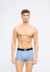 Άνδρας χωρίς μπλούζα με κοντά σγουρά μαλλιά, φοράει ανοιχτό μπλε αντρικά εσώρουχα με σκούρο λουρί μέσης που φέρει λευκό λογότυπο κροκόδειλου, στέκεται μπροστά σε απλό φόντο.