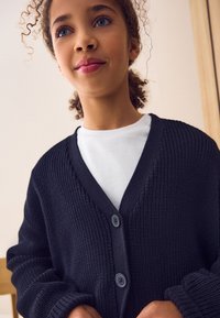 Mornarsko modra pletena jopica z V-izrezom in dvema gumbi, nošena čez belo srajco. Ima rebraste manšete in teksturirano tkanino po celotni površini.