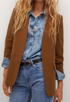 Kvinde iført en brun blazer over en denimskjorte med opkrogede ærmer, som er stoppet ned i højtaljede blå jeans med sort bælte og guldring på venstre hånd.