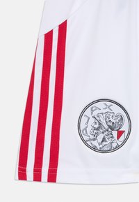 Weißes Sport-Trikot mit roten Streifenakzenten, einem runden Logo mit einer Figur und dem Text "AJAX" sowie einer glatten Textur.