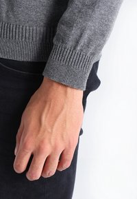 Grauer, gestrickter Pulloverärmel mit geripptem Muster, über einem dunklen Stoff getragen. Sichtbare Hand mit definierten Venen und Hautstruktur.