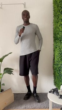 Pilka ilgomis rankovėmis marškiniai, juodos šortai, juodi batai, stovintis su mažuoju juodu įrenginiu. Netoliese ant balto stalo žalios augalas ir kamera.