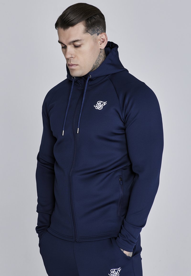 Námořnická mikina na zip vyrobená z elastického materiálu s vysokým límcem a kapucí na šňůrku, s bílým logem na hrudi a zipovými kapsami.