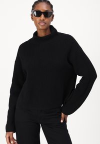 Maglione nero lavorato a maglia con colletto alto, texture a coste e maniche lunghe. Indossato con pantaloni neri e occhiali da sole.