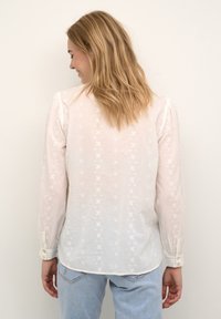 Kobieta z blond włosami do ramion, w białej haftowanej bluzce z długim rękawem i jasnoniebieskich dżinsach, stojąca przodem do białej ściany.