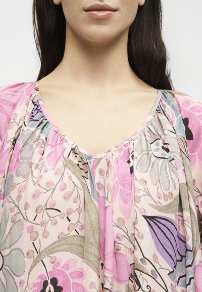 Γυναίκα με μακριά σκοτεινά μαλλιά, που φορά ροζ μπλούζα με λουλούδια, κοντό στρογγυλό λαιμόκοψη και πολύχρωμο βοτανικό σχέδιο.