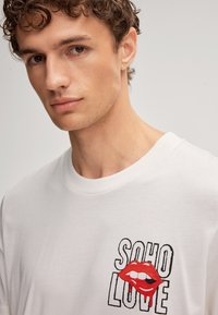 Jeune homme aux cheveux bouclés portant une chemise blanche avec un graphisme de lèvres rouges et le texte "SOHO LOVE" sur la poitrine.