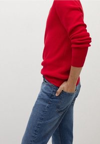 Roter gerippter Pullover, lange Ärmel, kombiniert mit blauen Jeans. Der Pullover hat eine figurbetonte Form, die Jeans sind gerade geschnitten und haben eine glatte Textur.
