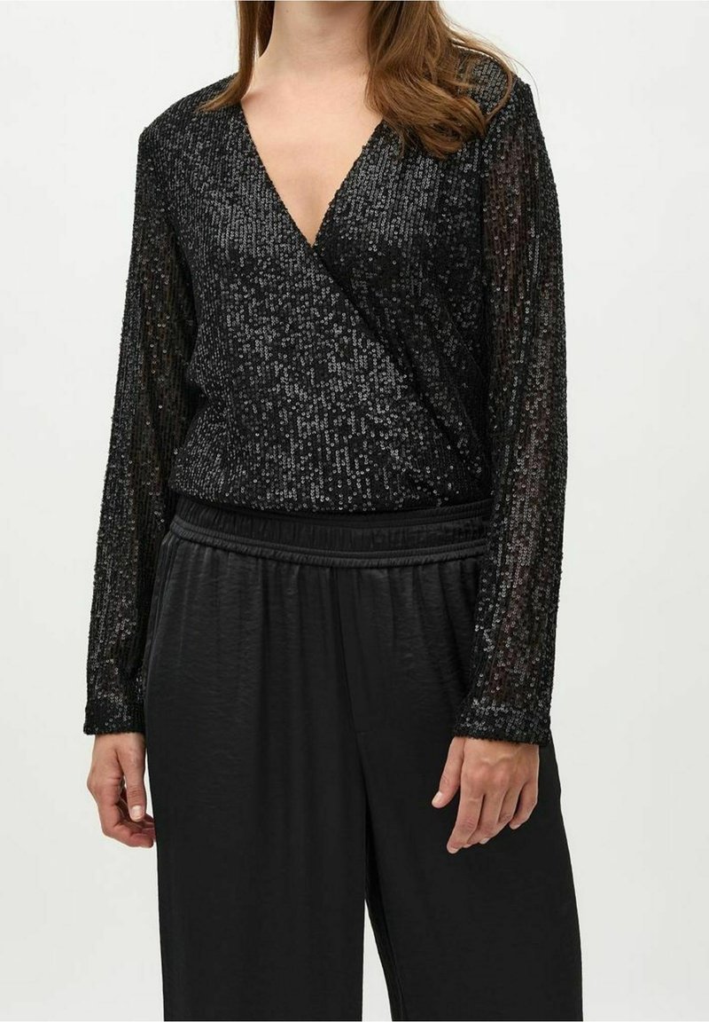 Femme portant un haut portefeuille noir à manches longues en sequins avec un décolleté en V profond et un pantalon large en satin noir sur fond uni.