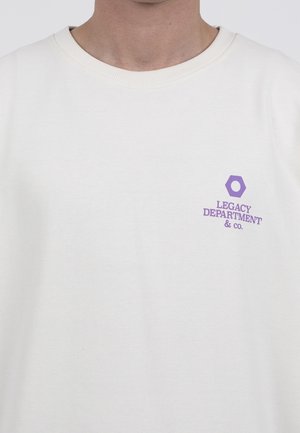 Osoba ubrana w prostą białą koszulkę z okrągłym dekoltem, z małym fioletowym sześciokątem i napisem „"Legacy Department & Co."” na lewej piersi.