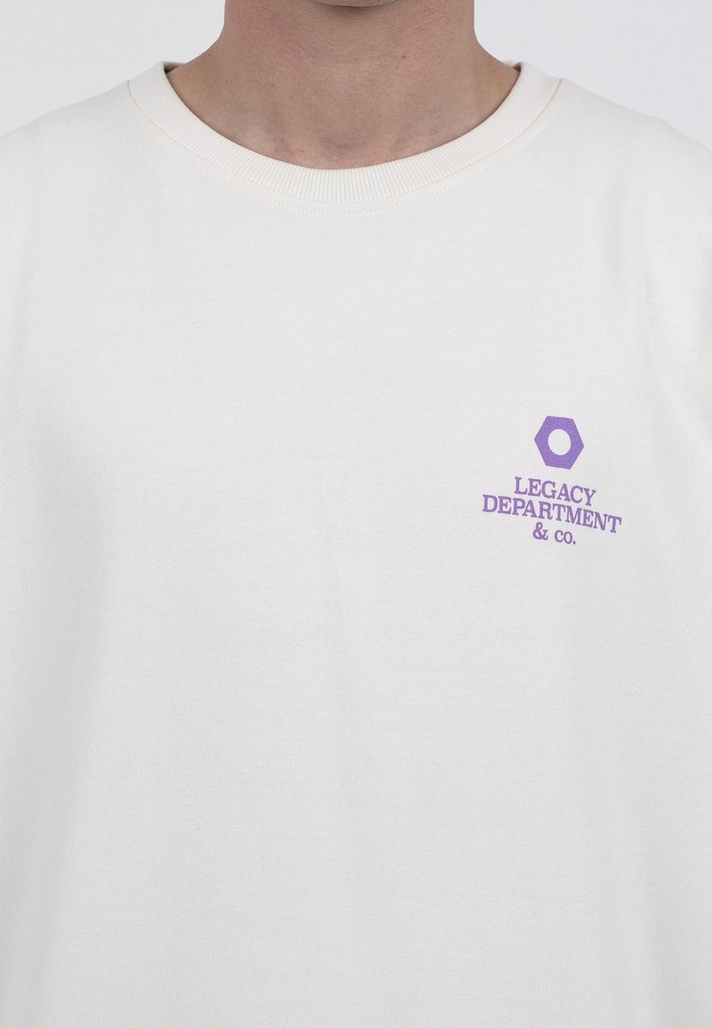 Personne portant un t-shirt blanc uni à col rond, avec un petit hexagone violet et le texte « "Legacy Department & Co." » sur la poitrine gauche.