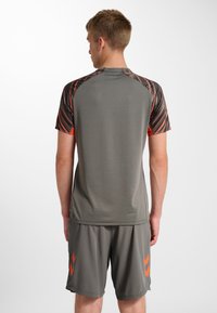 Jonge man staand met zijn rug naar de kijker, gekleed in een grijs sportshirt met zwart-oranje patroon op de mouwen en bijpassende grijze korte broek met oranje accenten.