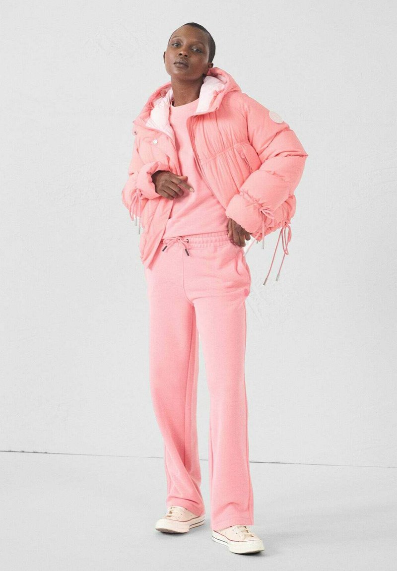 Giacca piumino rosa con dettagli a coulisse, abbinata a una felpa rosa coordinata e pantaloni della tuta. Completata con sneakers di colore chiaro.