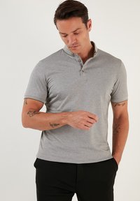 Světle šedé polo tričko s límečkem a zapínáním na tři knoflíky; má krátké rukávy a jemnou texturu. Nosené s tmavými kalhotami.