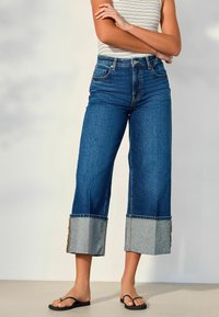 Persona con vaqueros de tiro alto y pierna ancha con grandes puños, camiseta sin mangas a rayas y chanclas negras, de pie con los brazos cruzados.