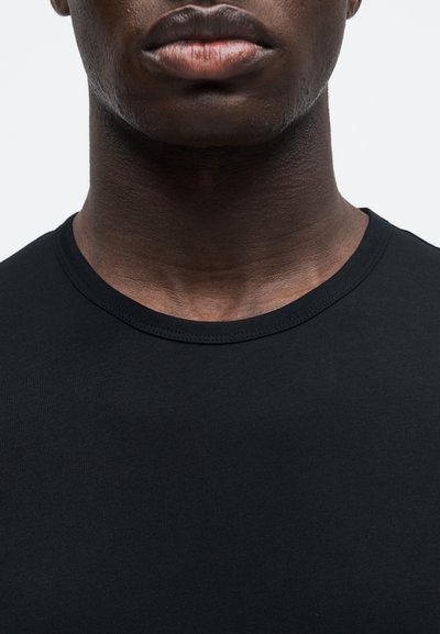 Gros plan sur la partie inférieure du visage et le cou d’une personne portant un t-shirt noir uni à col rond sur un fond clair uni.
