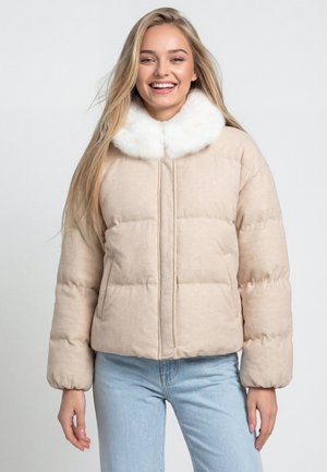Junge Frau lächelt, trägt eine beige gesteppte Jacke mit weißem Kunstpelzkragen und hellblaue Jeans, steht vor einem einfarbigen grauen Hintergrund.