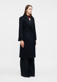 Γυναίκα που φοράει μακρύ μαύρο παλτό με ζώνη πάνω από παντελόνια με φαρδιά μπατζάκια και μπορντό κασκόλ, στέκεται μπροστά σε απλό ανοιχτό φόντο.