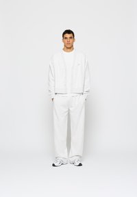 Fehér pamut kardigán gombokkal egy fehér póló felett, fehér nadrággal és világos sportcipőkkel párosítva. Egyszerű, letisztult dizájn és laza szabás.