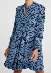 Kukkamekko, jossa tummansininen pohja ja sinisiä kukkakuvioita, pitkät hihat, napillinen kaulus ja röyhelöhelma. Valmistettu kevyestä kankaasta.