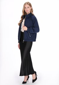 Marineblauw bomberjack met geplooide halslijn, ritszakken en elastische manchetten, gecombineerd met een lange zwarte satijnen rok en hoge hakken.
