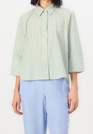Femme portant une chemise à boutons rayée verticale verte et blanche avec des manches trois-quarts et un pantalon bleu clair.