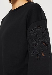 Felpa nera con colletto tondo, caratterizzata da un design a fiori intagliati testurizzati sulla manica sinistra. Tessuto morbido con una finitura liscia.