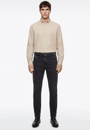 Mann trägt beige Hemd mit Knöpfen, schwarze Slim-Fit-Jeans und schwarze Lederstiefel, steht vor schlichtem weißem Hintergrund.