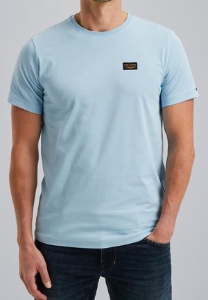 Mężczyzna w jasnoniebieskim t-shircie z krótkim rękawem z logo PME Legend na piersi, w połączeniu z ciemnoniebieskimi dżinsami, ręka w kieszeni.