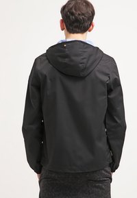 Personne vue de dos portant une veste à capuche noire et un pantalon sombre à motifs sur un fond clair uni.