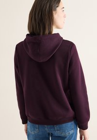 Bordeauxfarbener Kapuzenpullover mit weichem Material, gerippten Bündchen und Saum, von hinten gezeigt, kombiniert mit blauen Denim-Jeans.