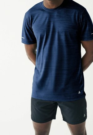 Mann, der ein dunkelblaues kurzärmliges Sportshirt und schwarze Shorts trägt, steht mit den Händen hinter dem Rücken vor einem schlichten hellen Hintergrund.