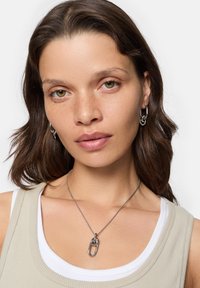 Sølv kæde halskæde med en overdimensioneret metallic sikkerhedsnål pendant, båret over en beige tank top og en hvid undertrøje. Glat, blank finish.