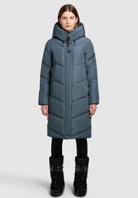 Gepolsterter, knielanger Mantel in dunklem Teal mit hohem Kragen und Kapuze. Verfügt über Chevron-Steppungen und Reißverschlusstaschen. Kombiniert mit schwarzen Stiefeln.
