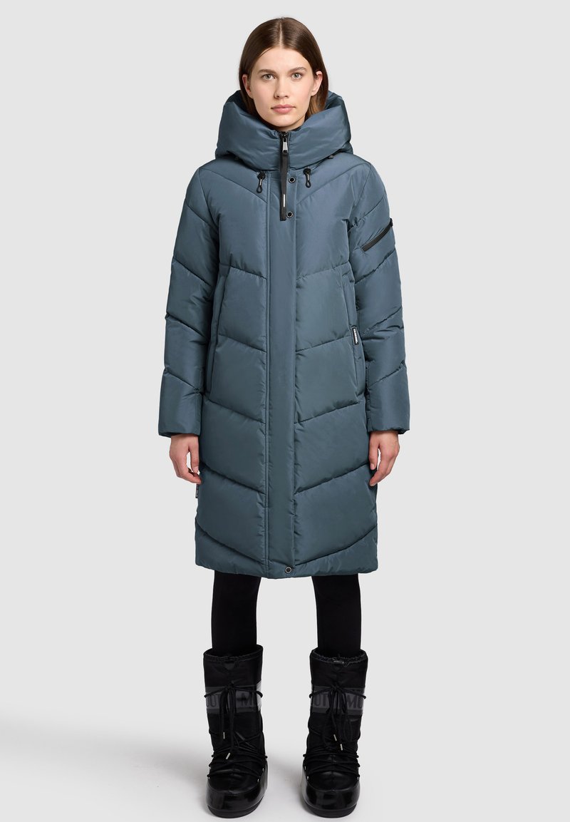 Gepolsterter, knielanger Mantel in dunklem Teal mit hohem Kragen und Kapuze. Verfügt über Chevron-Steppungen und Reißverschlusstaschen. Kombiniert mit schwarzen Stiefeln.