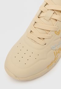 Beige joggesko laget av lær med en glatt tekstur. Har perforeringer og snøring, pyntet med dekorative håndskrevne detaljer i flere farger.