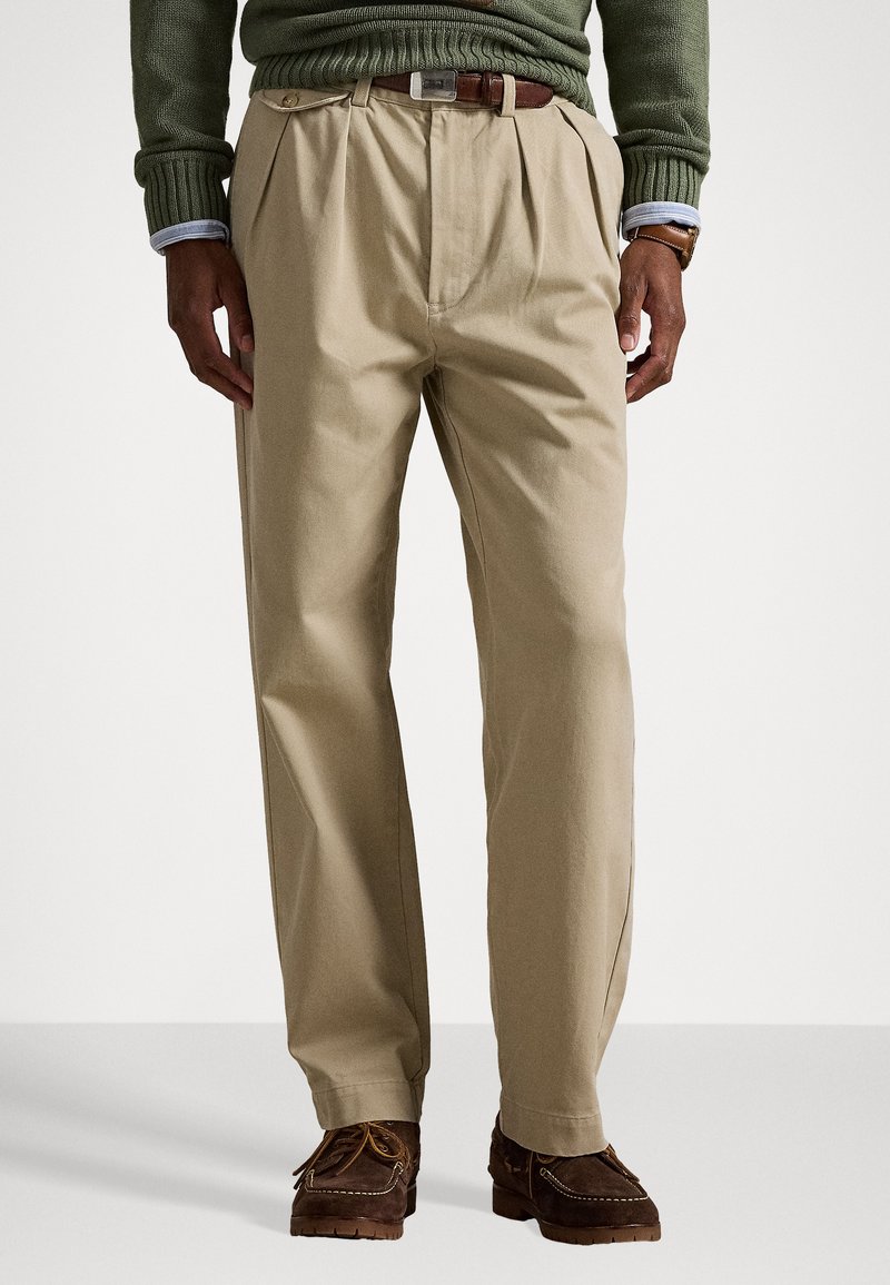 Beige rakott nadrág, könnyű anyagból, laza fazonban, vasalt gyűrődéssel; barna lábbelivel párosítva.