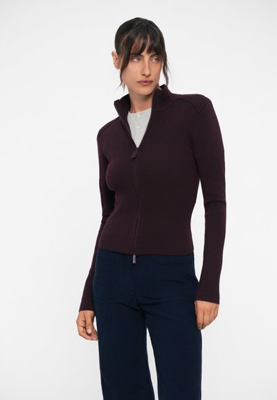 Burgundijas krāsas zip-up džemperis ar augstu apkakli, rievotām plecu šuvēm un garām piedurknēm. Uzvilkts virs pelēka henley krekla un saskaņots ar tumšām biksēm.