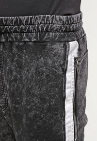 Šedé jogger kalhoty vyrobené z měkké látky se jemnou texturou; mají bílé boční pruhy a zapínací kapsu na pravém boku.