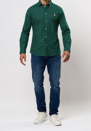 Mand iført en mørkegrøn skjorte med knapper, blå jeans og hvide sneakers, stående mod en ensartet lys baggrund.