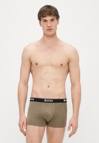 Ελαφρώς πράσινα μποξεράκια με μαύρη μέση που φέρει λευκό λογότυπο "BOSS". Ομαλή υφή και εφαρμοστή σχεδίαση, φορεμένα από έναν άνδρα μοντέλο.