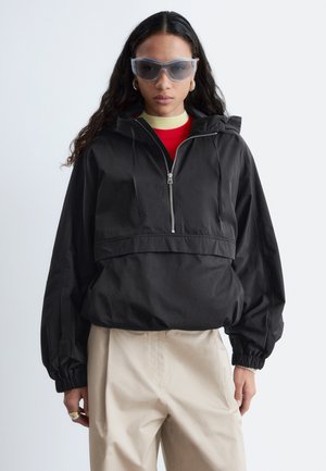 Kvinde iført oversized sort hættejakke med frontlomme, beige bukser, røde og gule lag-på-lag toppe og store grå solbriller.