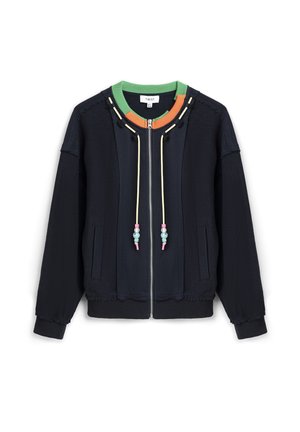 Marineblauwe jas met ritssluiting, groene en oranje kraag, crèmekleurige trekkoorden met kralen, zijzakken en geribbelde manchetten en zoom.