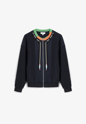 Marineblauwe jas met ritssluiting, groene en oranje kraag, crèmekleurige trekkoorden met kralen, zijzakken en geribbelde manchetten en zoom.