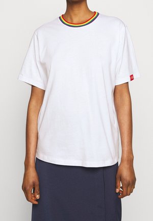 Persona que lleva una camiseta blanca de manga corta con cuello a rayas de arcoíris y una etiqueta roja con el logo en la manga, combinada con una falda oscura.