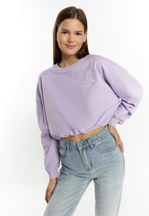 Sweatshirt corto di lavanda con maniche lunghe, scollatura rotonda e orlo elastico. Caratterizzato da testo ricamato bianco sul petto, abbinato a jeans azzurri chiari.