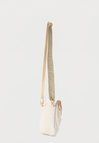 Μικρή μπεζ τσάντα χιαστί με ρυθμιζόμενο λουρί και μπροστινή τσέπη, κρεμασμένη μπροστά σε απλό λευκό φόντο.